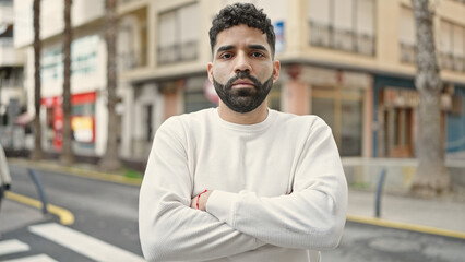 Young hispanic man standing with serious expression and arms crossed gesture at street