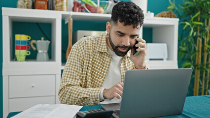 Young hispanic man talking on smartphone using laptop at dinning room