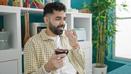 Young hispanic man drinking glass of wine sending voice message by smartphone at dinning room