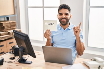 Hispanic man with beard working at the office with laptop holding thanks banner smiling happy...