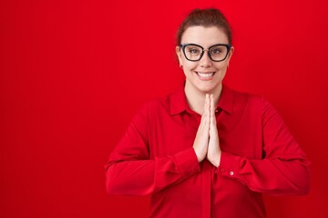 Young hispanic woman with red hair standing over red background praying with hands together asking for forgiveness smiling confident.