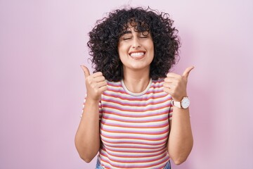 Young middle east woman standing over pink background success sign doing positive gesture with hand, thumbs up smiling and happy. cheerful expression and winner gesture.