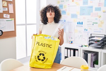 Young middle east woman holding recycling bag with plastic bottles at the office looking at the...