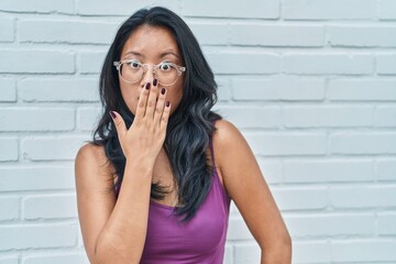 Asian young woman standing over bricks background covering mouth with hand, shocked and afraid for mistake. surprised expression