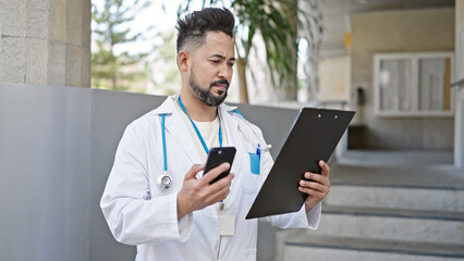 Young latin man doctor using smartphone reading document at hospital