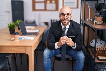 Young bald man business worker smiling confident sitting on table at office