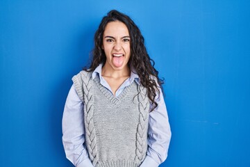 Young brunette woman standing over blue background sticking tongue out happy with funny expression....
