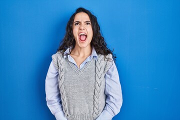 Young brunette woman standing over blue background angry and mad screaming frustrated and furious, shouting with anger. rage and aggressive concept.