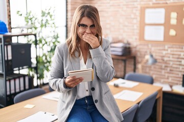 Young hispanic woman working at the office wearing glasses laughing and embarrassed giggle covering mouth with hands, gossip and scandal concept