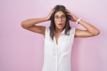 Brunette young woman standing over pink background wearing glasses crazy and scared with hands on head, afraid and surprised of shock with open mouth