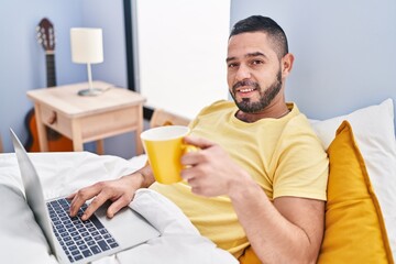 Young latin man using laptop drinking coffee at bedroom