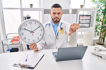 Hispanic doctor man holding clock at the clinic with angry face, negative sign showing dislike with...