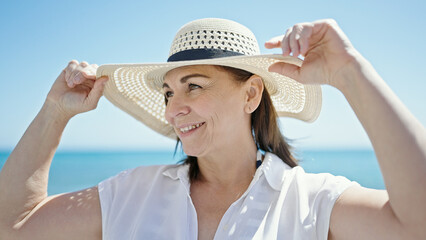 Middle age hispanic woman tourist smiling holding summer hat at the beach