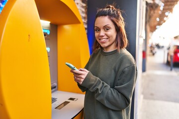 Young beautiful hispanic woman using smartphone standing by bank teller at street