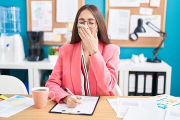 Young hispanic woman working at the office wearing glasses bored yawning tired covering mouth with...