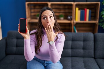Young brunette woman holding smartphone showing blank screen covering mouth with hand, shocked and...