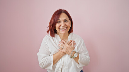 Middle age woman standing with hands on heart over isolated pink background