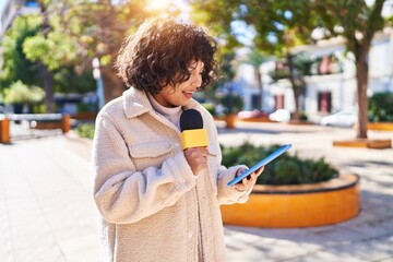 Young beautiful hispanic woman reporter working using microphone and touchpad at park