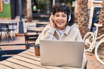 Young beautiful hispanic woman listening to music sitting on table at coffee shop terrace