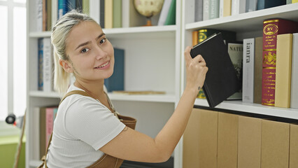 Young beautiful hispanic woman student smiling confident holding book of shelving at library university