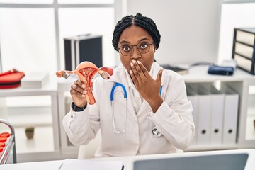 African american doctor woman holding anatomical model of female genital organ covering mouth with hand, shocked and afraid for mistake. surprised expression
