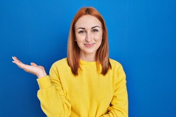 Young woman standing over blue background smiling cheerful presenting and pointing with palm of hand looking at the camera.
