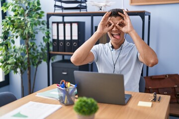 Young hispanic man working at the office wearing headphones doing ok gesture like binoculars...