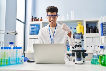 Young hispanic man working at scientist laboratory doing video call smiling happy pointing with hand and finger