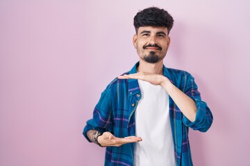 Young hispanic man with beard standing over pink background gesturing with hands showing big and large size sign, measure symbol. smiling looking at the camera. measuring concept.