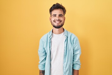 Young hispanic man with tattoos standing over yellow background with a happy and cool smile on...