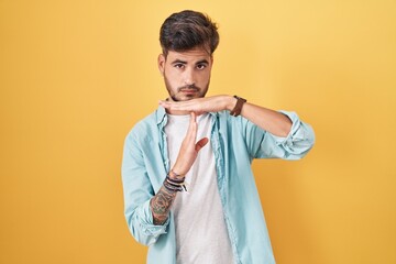 Young hispanic man with tattoos standing over yellow background doing time out gesture with hands,...