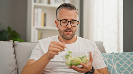 Grey-haired man eating salad sitting on the sofa at home