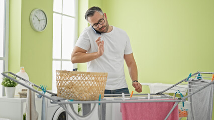 Grey-haired man talking on smartphone hanging clothes on clothesline at laundry room