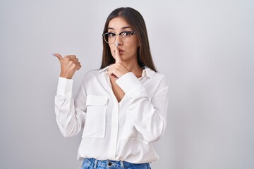 Young brunette woman wearing glasses asking to be quiet with finger on lips pointing with hand to the side. silence and secret concept.