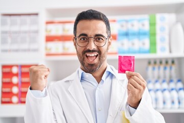 Hispanic man with beard working at pharmacy drugstore holding condom screaming proud, celebrating victory and success very excited with raised arms