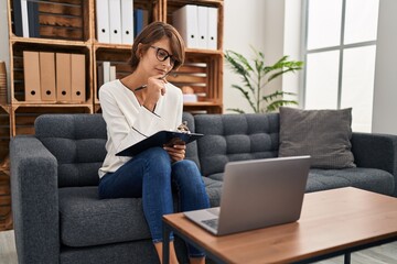 Young beautiful hispanic woman psychologist writing on document using laptop at psychology clinic