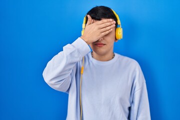 Non binary person listening to music using headphones covering eyes with hand, looking serious and sad. sightless, hiding and rejection concept