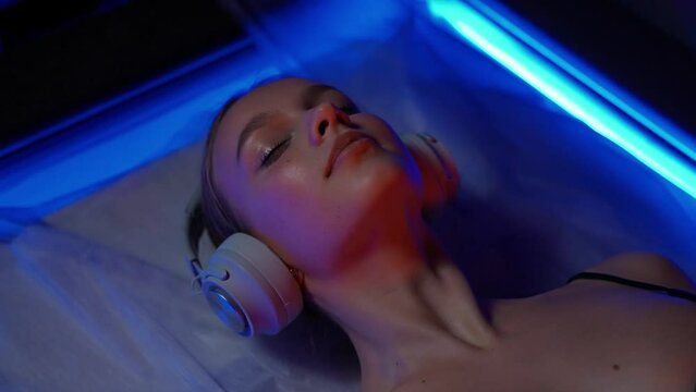 Young woman with headphones lies down in the sensory deprivation chamber, neon light, relax in spa, portrait.