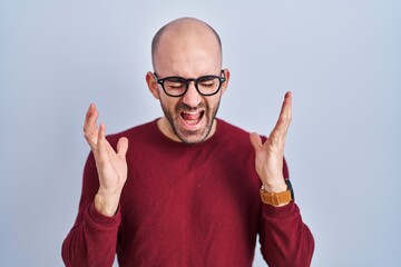 Naklejka premium Young bald man with beard standing over white background wearing glasses celebrating mad and crazy for success with arms raised and closed eyes screaming excited. winner concept