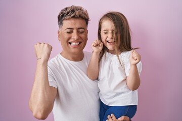 Young father hugging daughter over pink background screaming proud, celebrating victory and success very excited with raised arms