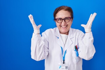 Senior woman with glasses wearing scientist uniform celebrating mad and crazy for success with arms raised and closed eyes screaming excited. winner concept