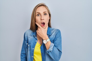 Young blonde woman standing over blue background looking fascinated with disbelief, surprise and...