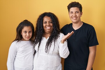 Family of mother, daughter and son standing over yellow background smiling cheerful presenting and pointing with palm of hand looking at the camera.