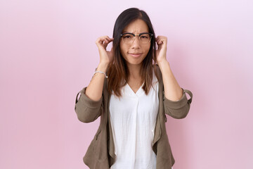 Middle age chinese woman wearing glasses over pink background covering ears with fingers with annoyed expression for the noise of loud music. deaf concept.