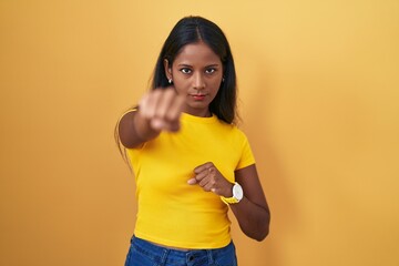 Young indian woman standing over yellow background punching fist to fight, aggressive and angry attack, threat and violence