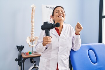Middle age hispanic doctor woman holding therapy massage gun at wellness center screaming proud, celebrating victory and success very excited with raised arm