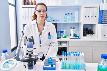 Fototapeta premium Young hispanic woman working at scientist laboratory relaxed with serious expression on face. simple and natural looking at the camera.