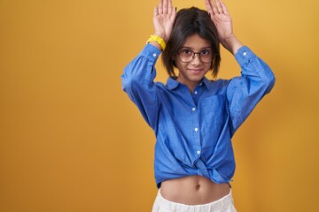 Young girl standing over yellow background doing bunny ears gesture with hands palms looking...