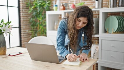 Young beautiful hispanic woman using laptop writing notes at dinning room