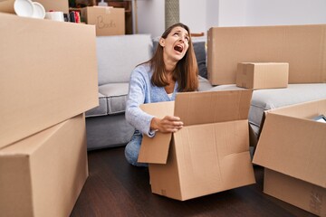 Young woman moving to a new home looking inside cardboard box angry and mad screaming frustrated and furious, shouting with anger looking up.